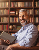 Mann mit Bart und Buch in der Hand lächelt, Bücherregal im Hintergrund (KI-generiertes Bild)