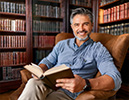 Mann mit Buch in der Hand sitzt vor einem Bücherregal und lächelt (KI-generiertes Bild)